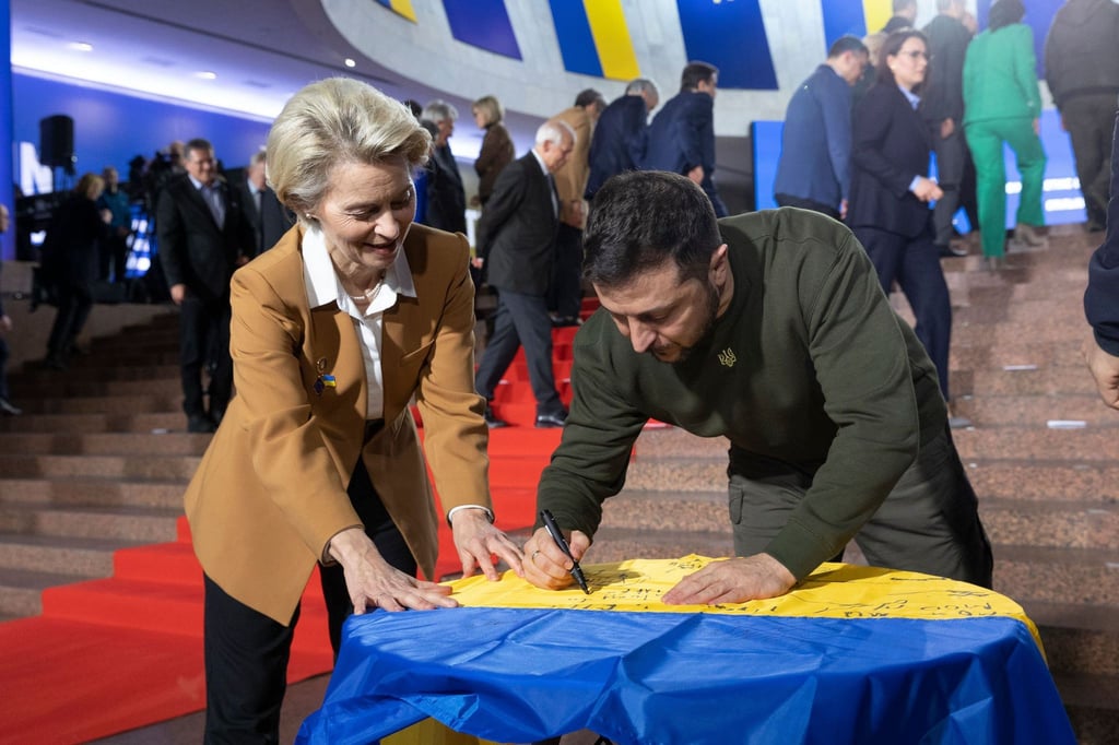 Wolodymyr Selenskyj und Ursula von der Leyen schreiben ihre Wünsche auf eine ukrainische Flagge.