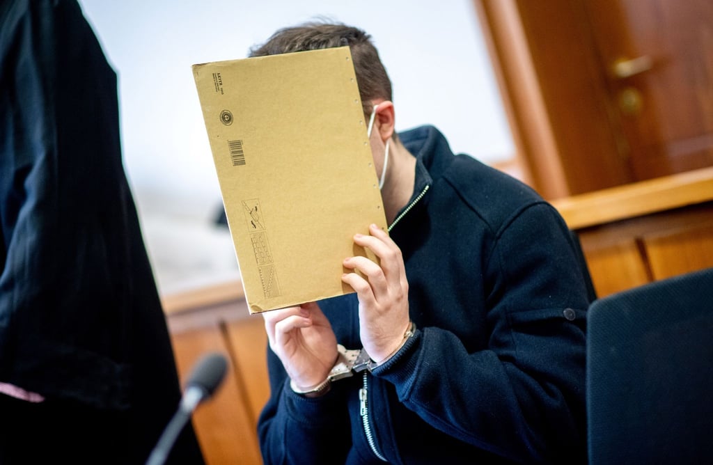 Der Angeklagte sitzt im Gerichtssaal und hält sich einen Aktendeckel vor das Gesicht.
