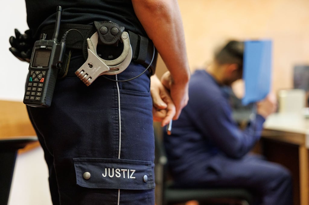 Ein Justizbeamter, mit Funkgerät und Handschellen, steht neben einem Angeklagten im Landgericht.