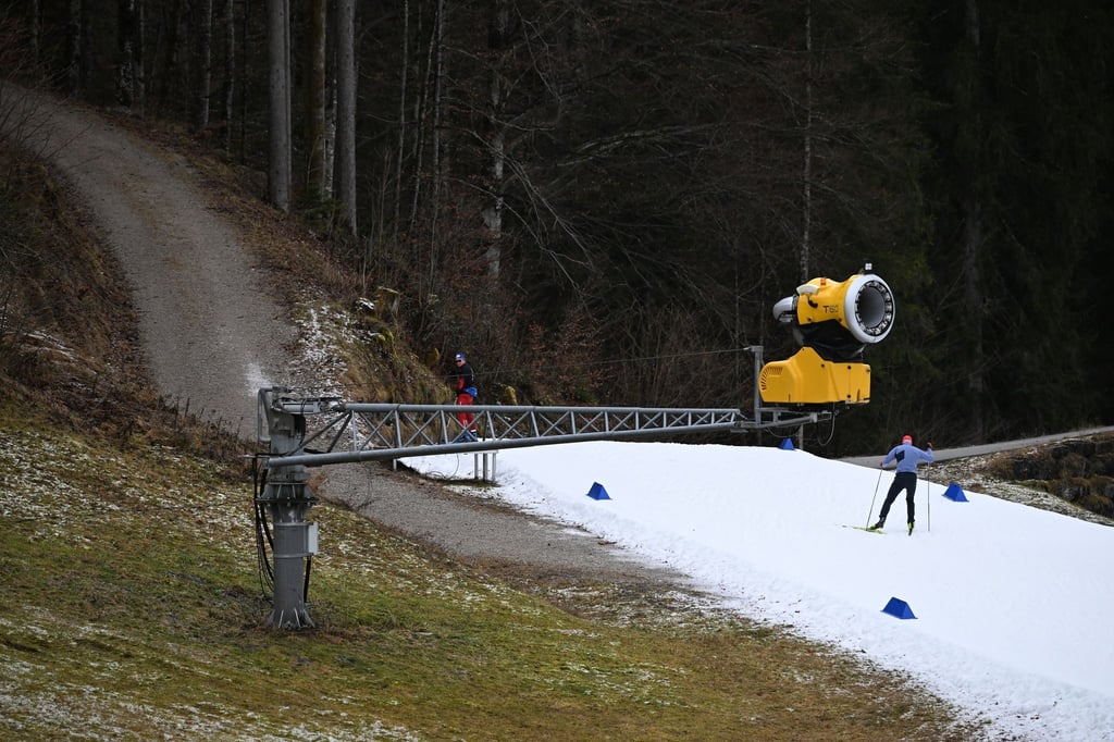 Eine Schneekanone ist an der Loipe aus Kunstschnee zu sehen, während abseits kein Schnee liegt.
