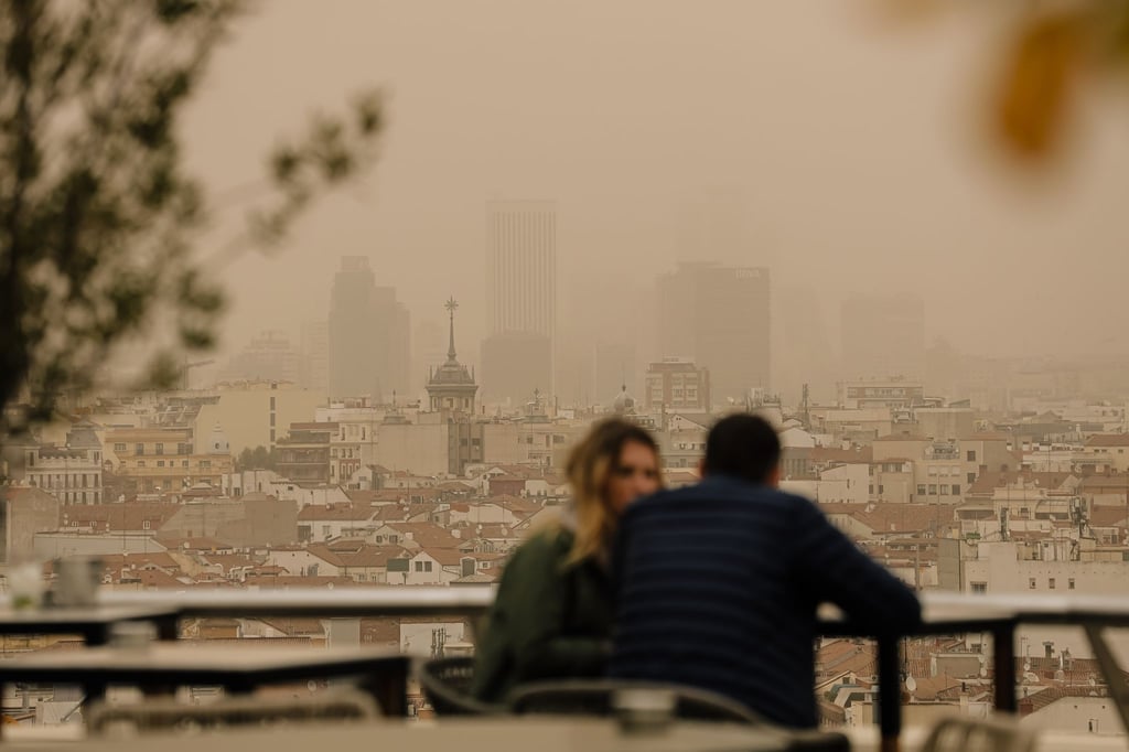Zwei Menschen sitzen in Madrid auf einer Terrasse, während eine Wolke mit Sahara-Staub über die spanische Hauptstadt zieht. Die typische Auswirkung von Sahara-Staub ist ein roter oder orangener Himmel (Archivbild).