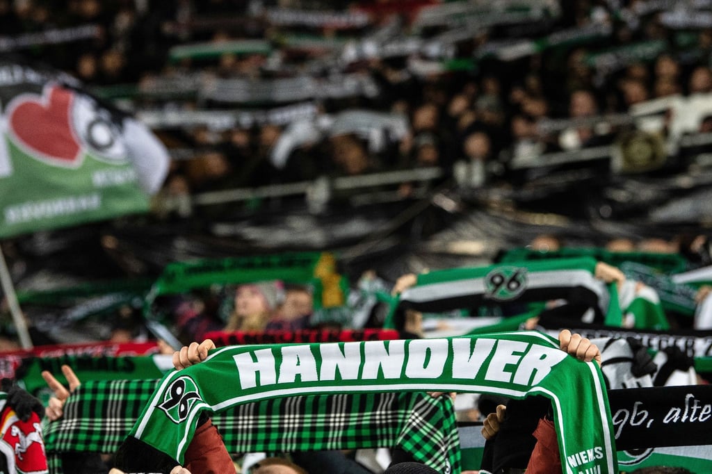 Fans von Hannover 96 stehen mit ihren Schals auf der Tribüne.