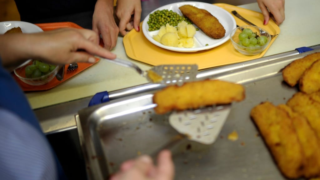 Eine Küchenhelferin legt an einer Gesamtschule ein Stück Fisch auf den Teller eines Schülers.