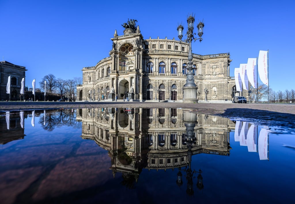 Die Semperoper spiegelt sich bei Sonnenschein in einer Pfütze auf dem Theaterplatz.