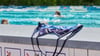 Ein Badeanzug liegt auf einer Mauer, dahinter schwimmen Menschen im Becken ihre Bahnen.
