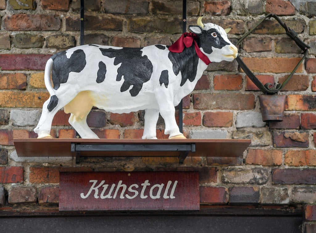 Eine kleine Kuh aus Plastik steht auf einem Brett über einer Tür mit der Aufschrift „Kuhstall“.