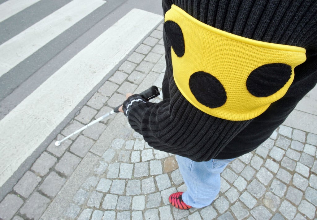 Ein blinder Mann geht mit seinem weißen Taststock und einer Armbinde mit drei Punkten über eine Straße in Hannover.
