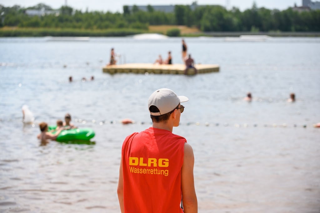 Ein Mitglied der Deutschen Lebens-Rettungs-Gesellschaft (DLRG) steht in einem Strandbad am Ufer.