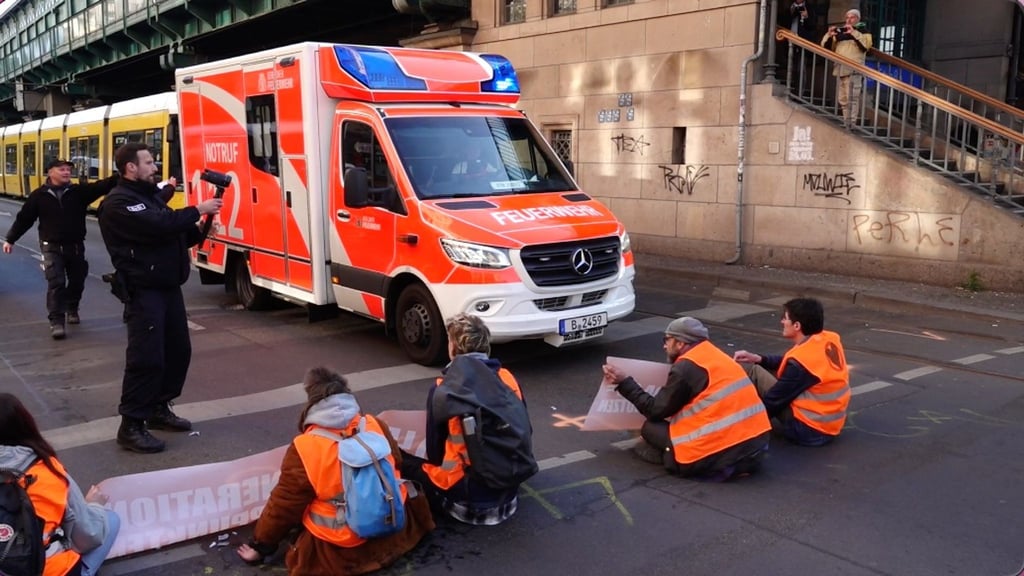 Aktivisten der Gruppe Letzte Generation blockieren die Fahrbahn an der Schönhauser Allee, Ecke Eberswalder Str., während ein Rettungswagen zum Einsatz unterwegs ist und daran vorbeifährt.