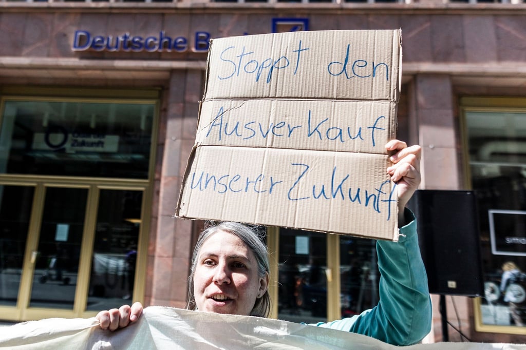 Demonstrantin mit Schild „Stoppt den Ausverkauf unserer Zukunft“ vor einer Deutschen-Bank-Filiale.