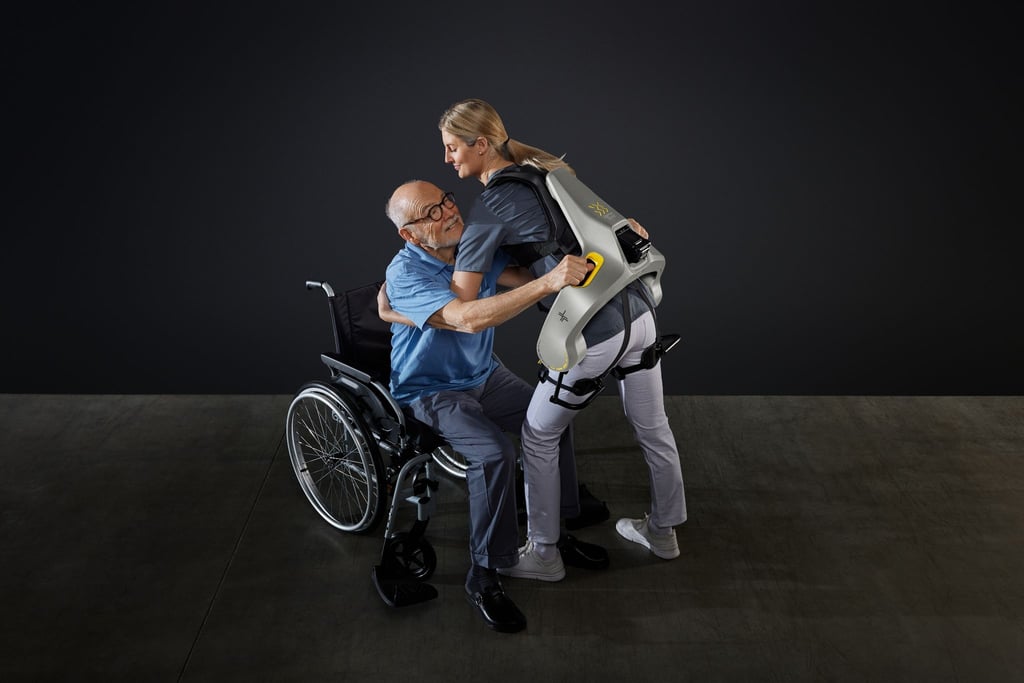 Eine Frau hebt mit Hilfe eines intelligenten Kraftanzugs der Firma German Bionic einen Mann aus einem Rollstuhl.