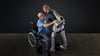 Eine Frau hebt mit Hilfe eines intelligenten Kraftanzugs der Firma German Bionic einen Mann aus einem Rollstuhl.