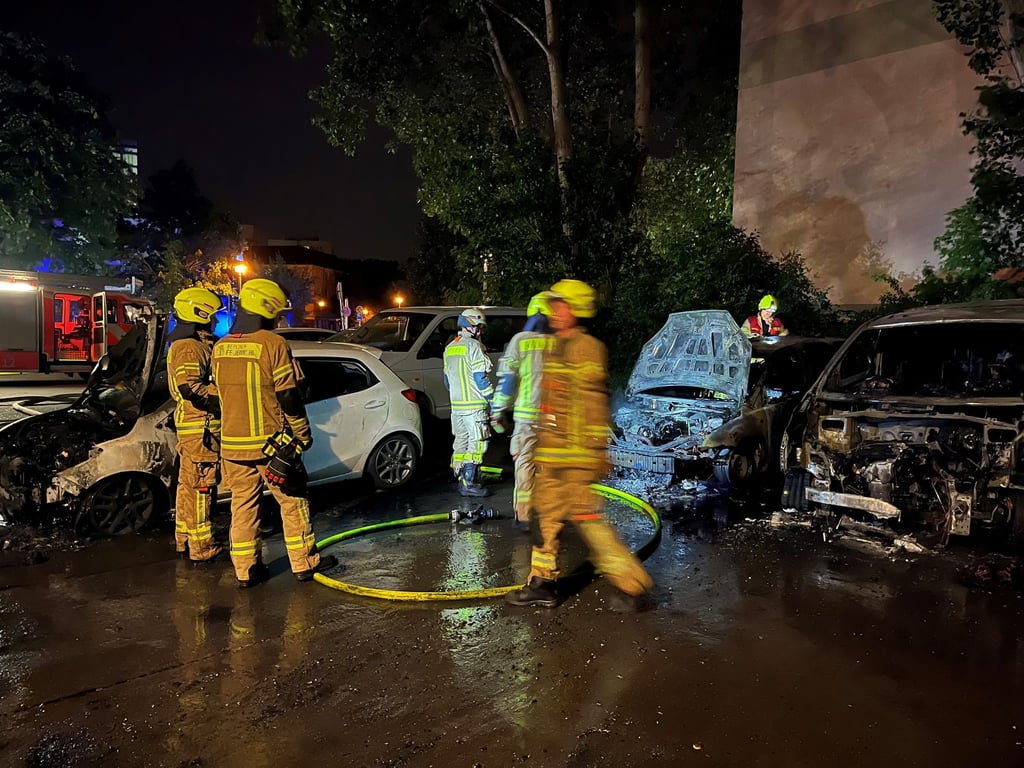 Feuerwehrleute sind auf einem Parkplatz im Einsatz, auf dem drei Fahrzeuge abgebrannt sind.