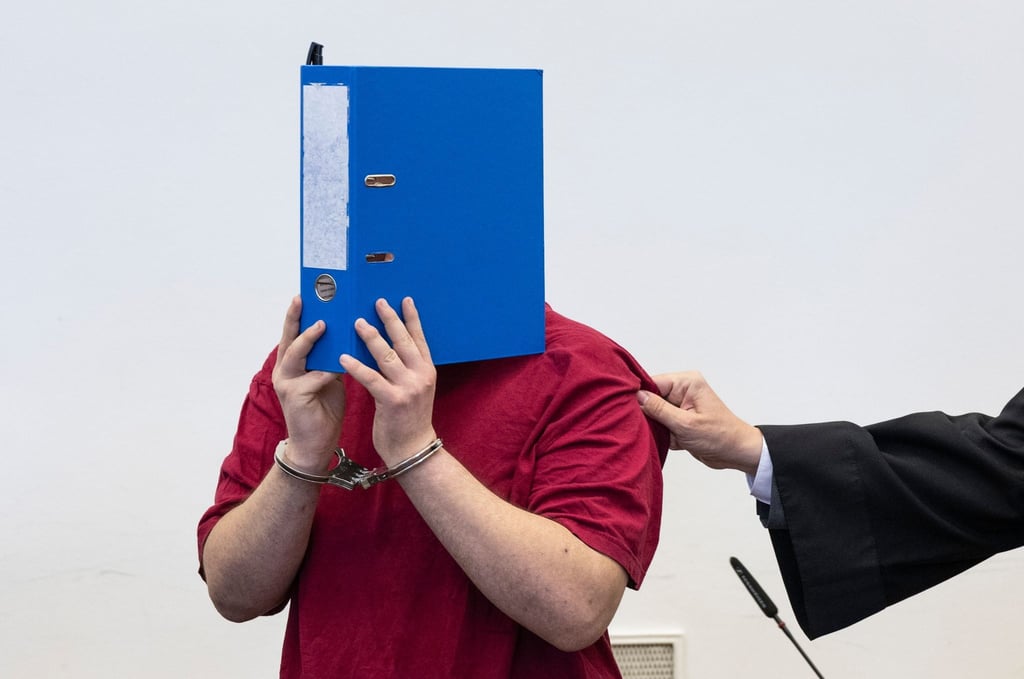 Der Angeklagte steht hinter der Anklagebank und verdeckt sein Gesicht hinter einem Aktenordner.