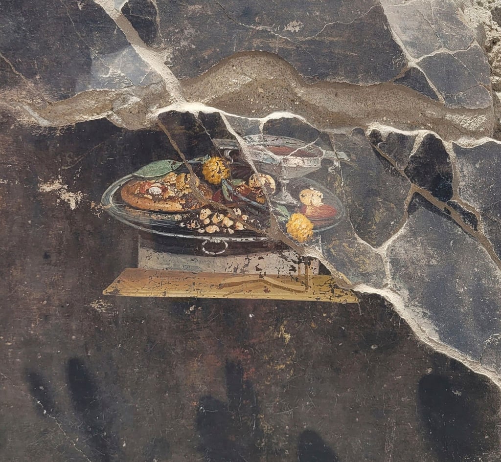 Die Wand eines pompejanischen Hauses mit einem Fresko, das einen Tisch mit Speisen darstellt, darunter auch eine Art Pizza.
