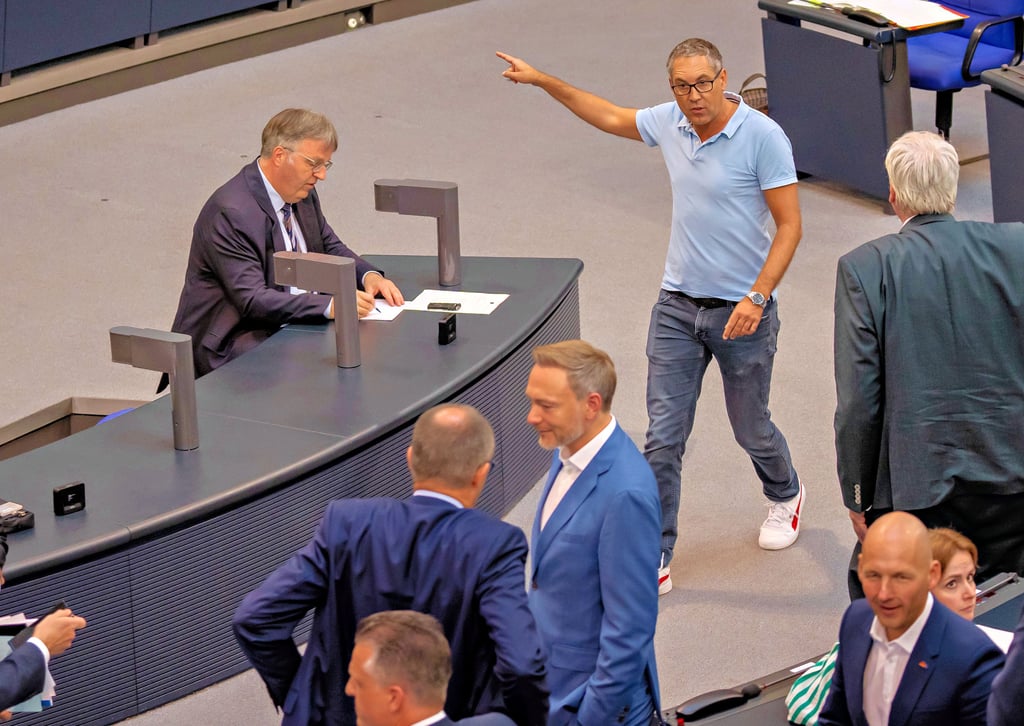SPD-Bundestagsabgeordneter Michael Schrodi läuft gestern im hellblauen Polohemd pöbelnd durch den Bundestag. Im Vordergrund steht Bundeswirtschaftsminister Christian Lindner (FDP) im blauen Anzug, links daneben CDU-Chef Friedrich Merz.