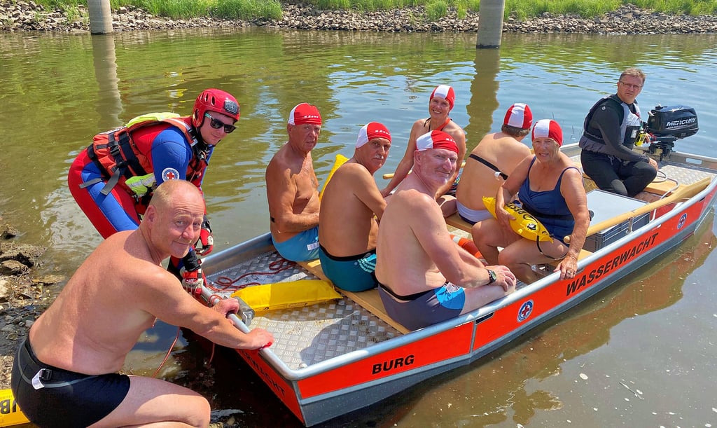Sechs Schwimmer sitzen im Boot der Wasserwacht und können es nicht erwarten, zu schwimmen. In Kürze werden sie in Richtung Autobahnbrücke gebracht, von wo aus sie zurückschwimmen müssen.