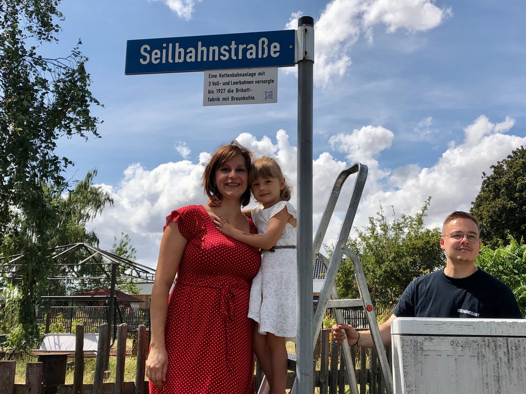 Ärztin Claudia Günther und ihre Tochter Frieda enthüllen im Müchelner Ortsteil Stöbnitz das Erklärschild für den Namen Seilbahnstraße. Ordnungsamtsmitarbeiter Sebastian Röhl hilft dabei.
