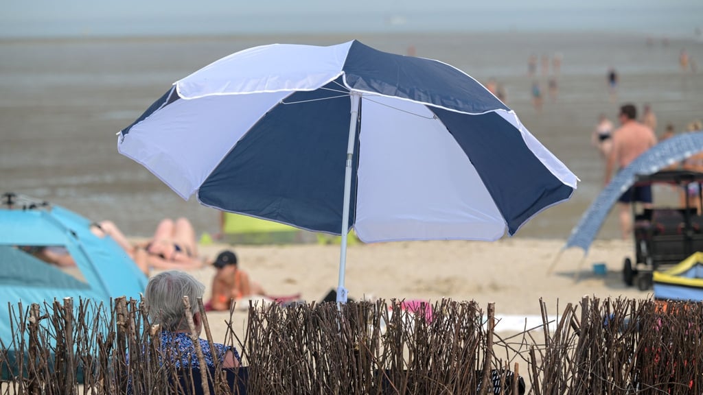 Eine ältere Frau sitzt sommerlichem am Badestrand unter ihrem Sonnenschirm.
