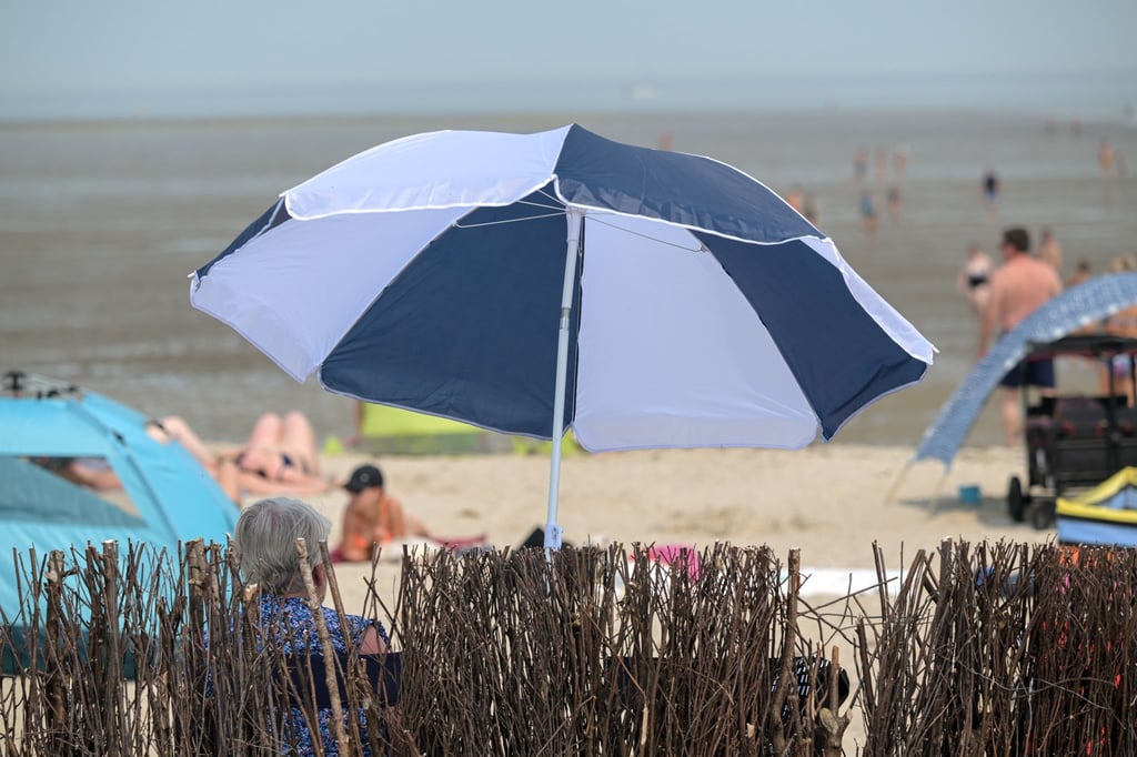Eine ältere Frau sitzt sommerlichem am Badestrand unter ihrem Sonnenschirm.