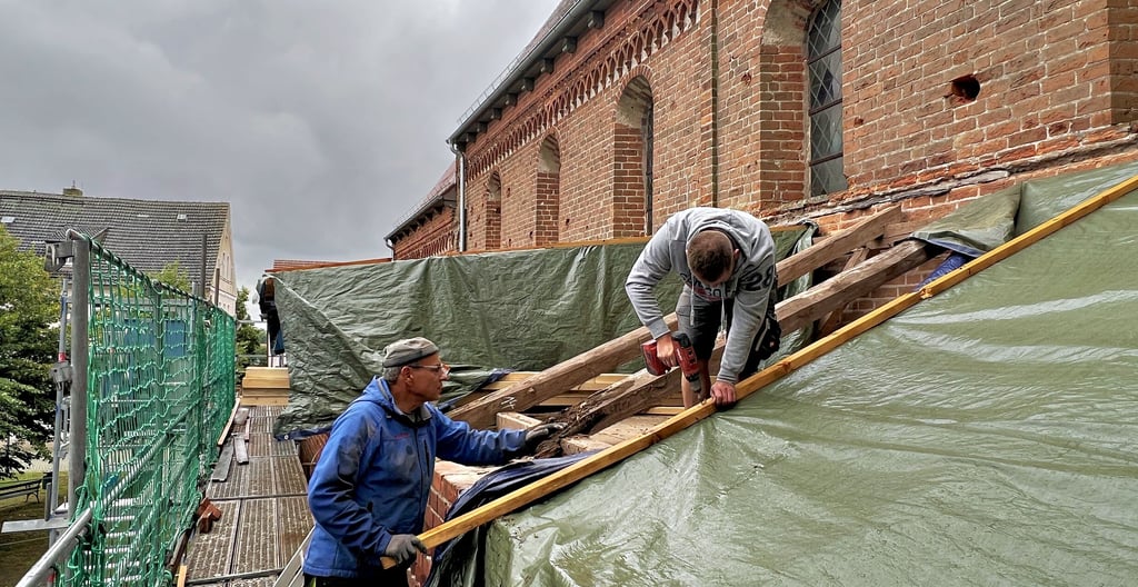 Die Zimmerleute aus dem brandenburgischen Herzberg sind mit dem Dachgebälk der Sandauer Kirche schon gut vertraut. Sie errichteten zuerst ein Wetterschutzdach aus großen Planen, das nach dem Abriss der Dachpappe zum Auswechseln des Gebälks aufgeständert wurde – wie im Hintergrund zu sehen.