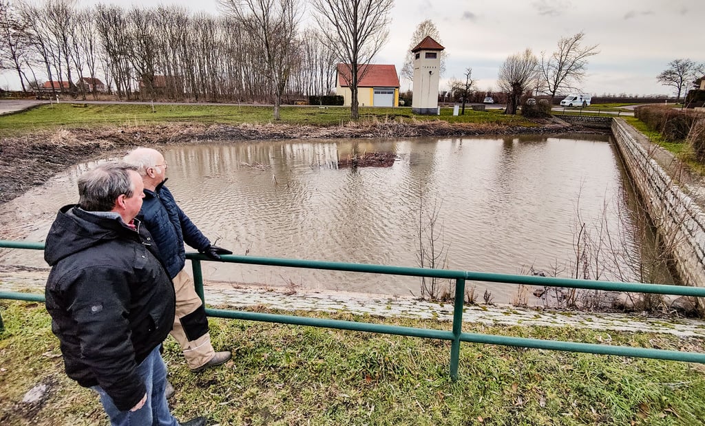 Ortsbürgermeister Michael Winter (von links) und Roland Schumann aus dem Ortschaftsrat (beide Bürgerliste) stehen am Dorfteich in Bothfeld und blicken auf die übrig gebliebene Mittelinsel, die inzwischen vom Regen umspült wurde.