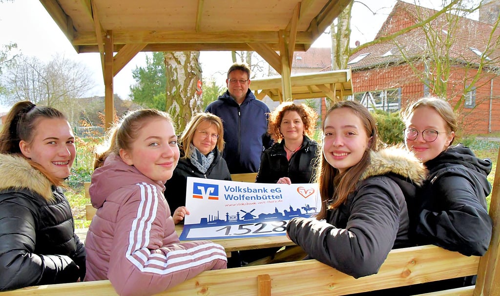 Fiona Baier (vorn; v. l.), Lucy Goldacker, Leonie Herrmann und Angelina Hartmann sitzen zur Probe auf den neuen überdachten Bänken auf einem der beiden  Pausenhöfe der Calvörder Sekundarschule „Brüder Grimm“. Kevyne Wendt (hinten; l.) von der Volksbank Wolfenbüttel überreicht den Scheck in Höhe von 1.528 Euro an Marén Gericke vom Schulförderverein und an Schulleiter Claudio Kühn.