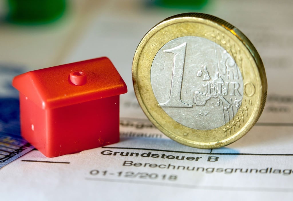 Eine Euro-Geldmünze sowie ein Spielzeughaus auf einem Abgabenbescheid.