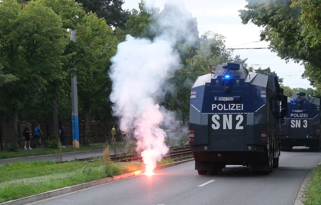 Pyrotechnik zündet auf einer Straße, während Wasserwerfer der Polizei vorüberfahren.