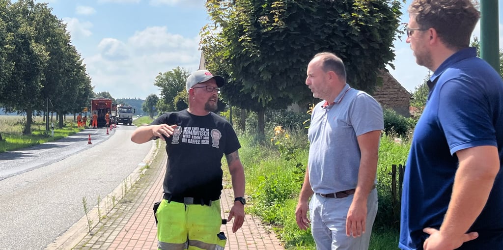 Polier Daniel Thieme (Kutter Spezialstraßenbau) im Gespräch mit Thomas Staudt und Marco Radke (rechts). Sie lassen sich vor Ort erläutern, wie weit die Arbeiten fortgeschritten sind. Im Hintergrund die große Maschine, die den Belag aufbringt.