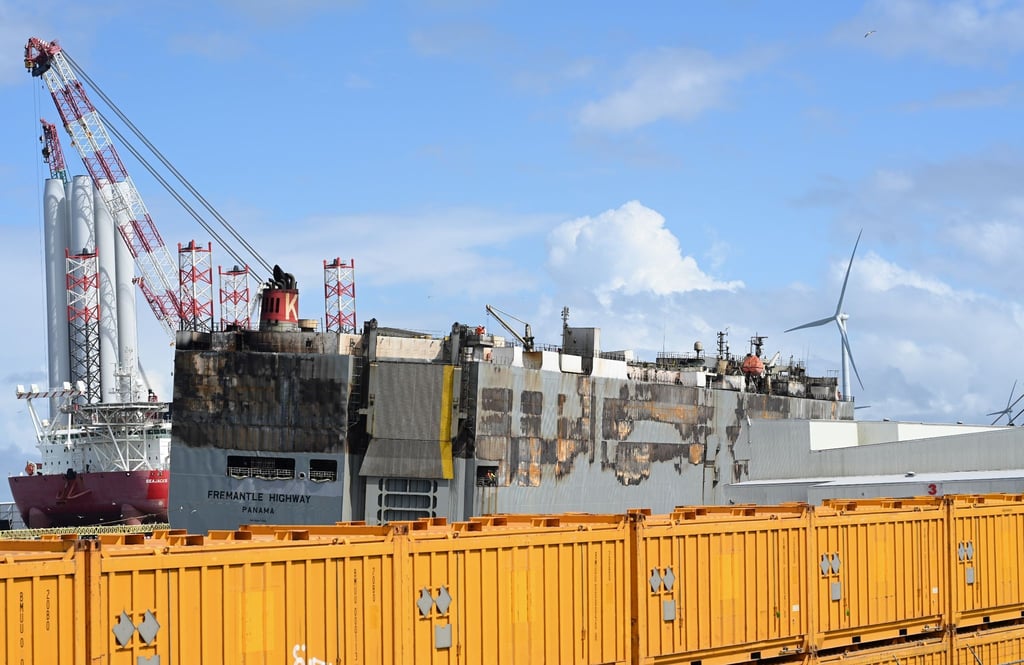 Der schwer beschädigte Autofrachter “Fremantle Highway„ liegt im Hafen, abgeschirmt durch gelbe Container.