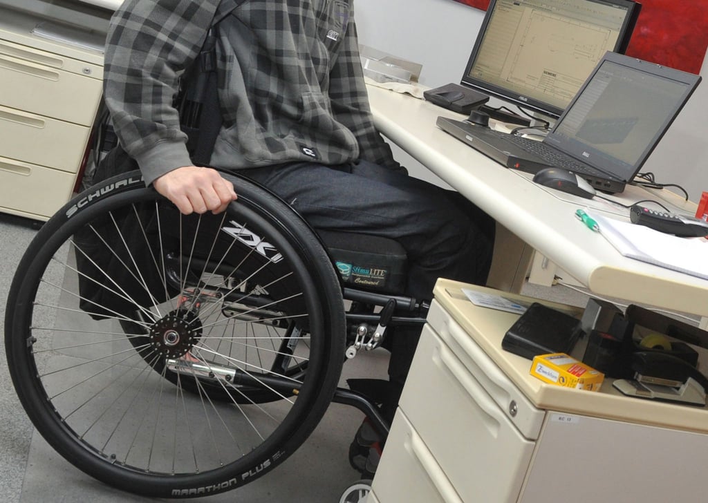 Ein Mann sitzt in einem Rollstuhl an seinem Arbeitsplatz am Schreibtisch.