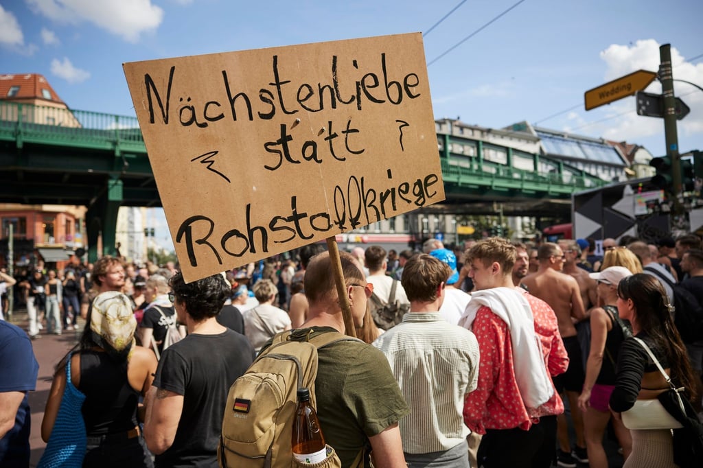 Ein Feiernder trägt ein Schild mit der Aufschrift „Nächstenliebe statt Rohstoffkriege“ bei der Technoparade Zug der Liebe durch Berlin.