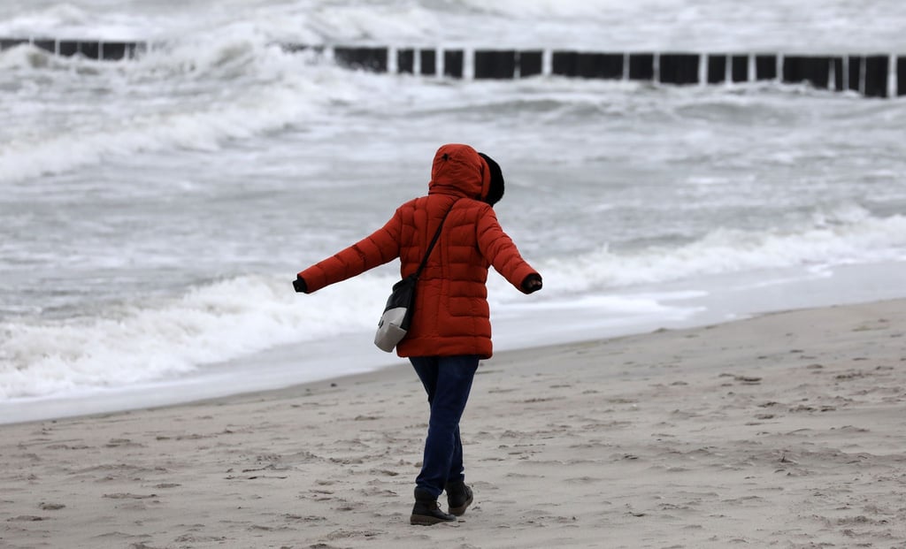 Eine Spaziergängerin geht bei Wind und Regen über einen Strand.
