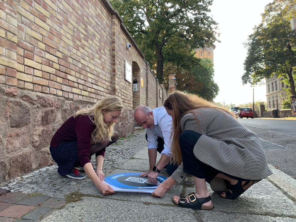 Anna Petersen, Mitglied der Chefredaktion (links), Johanna Huber vom Landesnetzwerk der Migrantenorganisationen (rechts) und MZ-Redakteur Dirk Skrzypczak bringen vor der Synagoge in Halle den ersten Aufkleber mit dem QR-Code zum aktuellen Artikel samt Video auf.