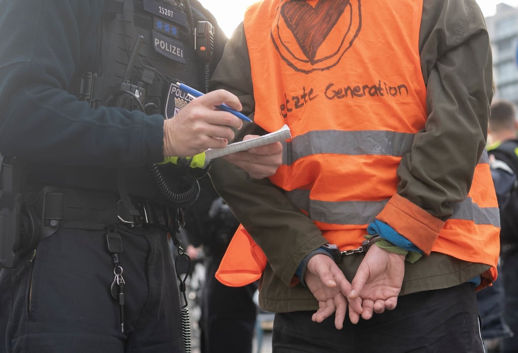 Ein Aktivist der Klimaschutz-Gruppe Letzte Generation steht in Handschellen neben einem Polizeibeamten.