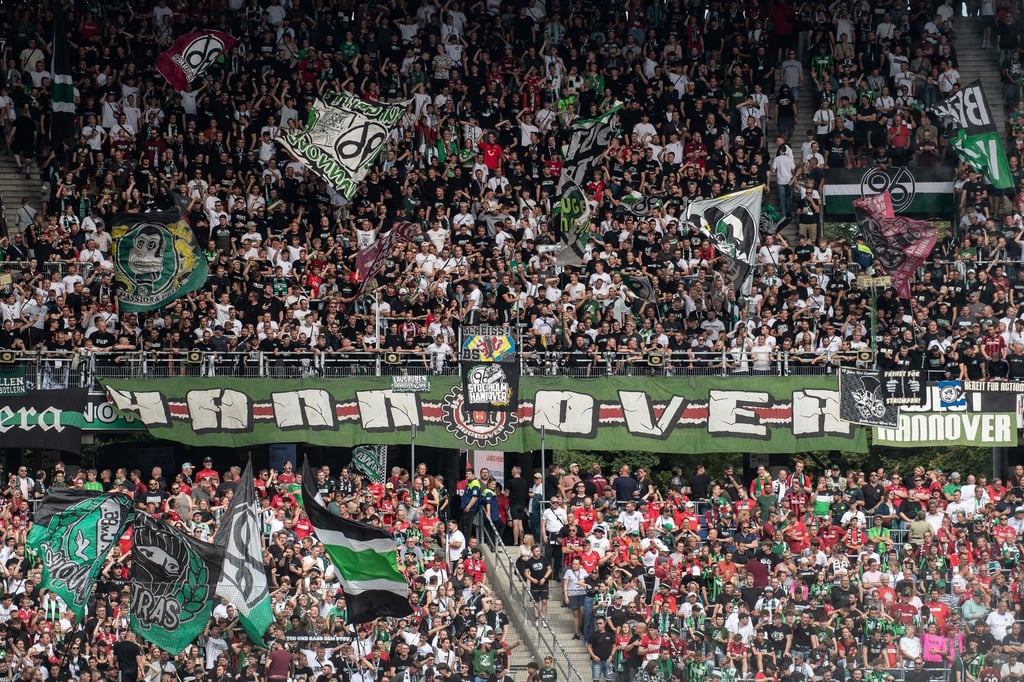 Fans von Hannover 96 stehen mit Fahnen auf der Tribüne.