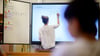 Eine Lehrerin schreibt eine Mathematikaufgabe auf eine digitale Schultafel im Klassenraum.