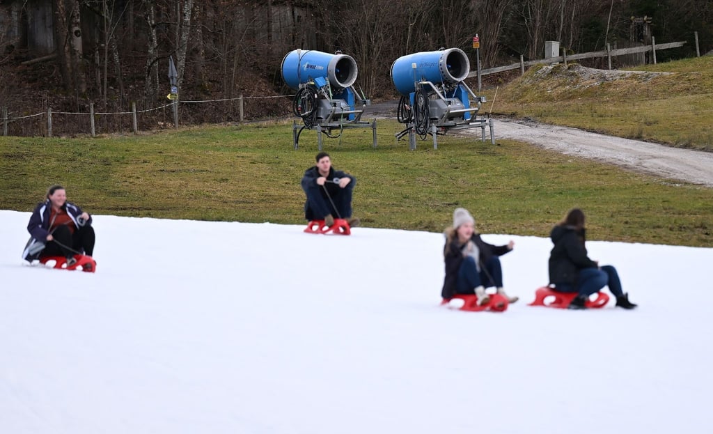 Rodler fahren auf einer Piste an im Grünen stehenden Schneekanonen vorbei.