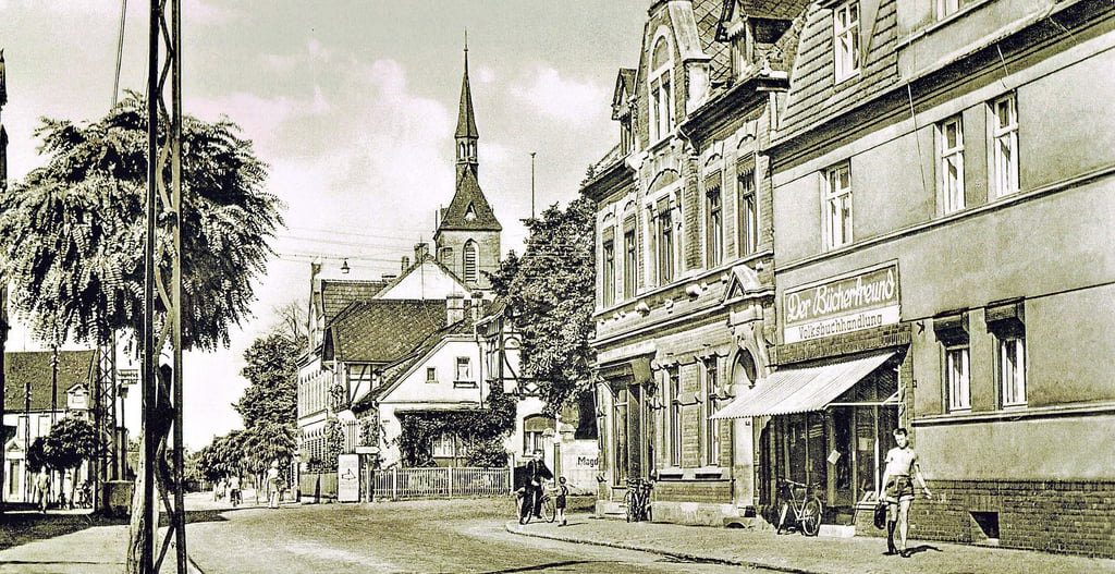 Die Martin-Schwantes-Straße in Gommern in den 1960er Jahren. Schon Jahrzehnte zuvor war sie eine beliebte Einkaufsstraße in Gommern, wie man aus dem Leserbrief aus dem Jahre 1903 erfahren kann. Vorn rechts sieht man die ehemalige Buchhandlung „Der Bücherfreund“, hinter dem markanten Fachwerkhaus in der Mitte des Bildes das ehemalge Postamt sowie die katholische Kirche „Herz Jesu“.