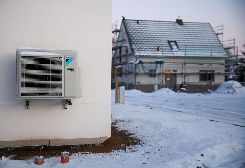 Eine Luftwärmepumpe hängt bei Schnee und Eis an einer Hauswand in einem neu entstehenden Einfamilienhausgebiet.