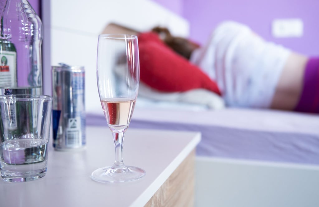 Eine Frau liegt in ihrem Bett, neben dem Flaschen und Gläser mit Alkohol stehen.