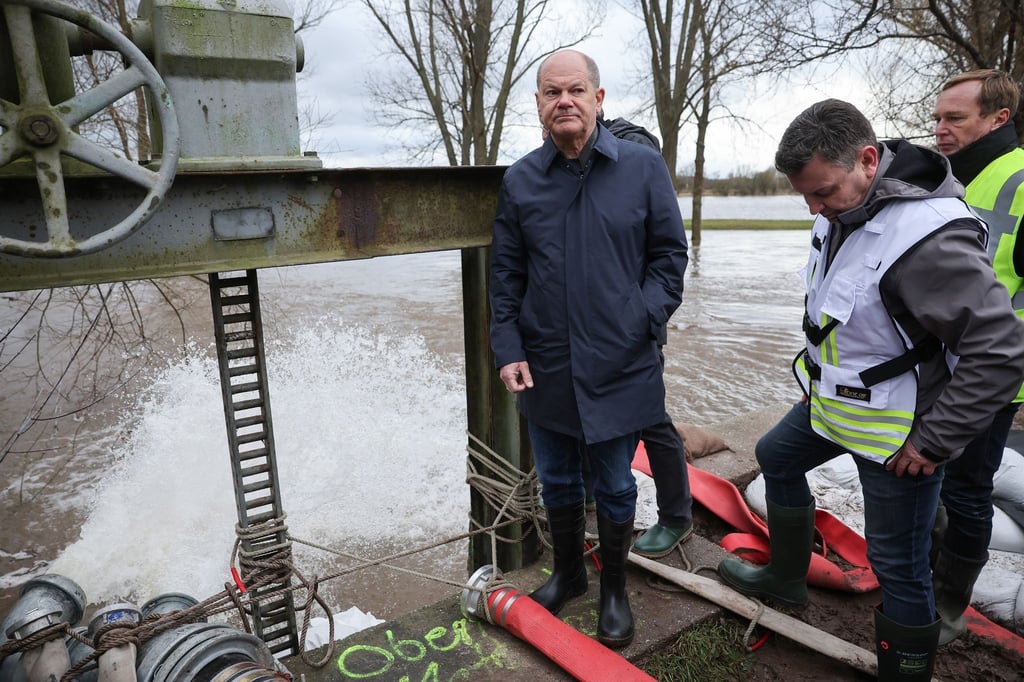 Bundeskanzler Olaf Scholz steht an einem Wehr, an dem Schläuche abgepumptes Wasser abführen.