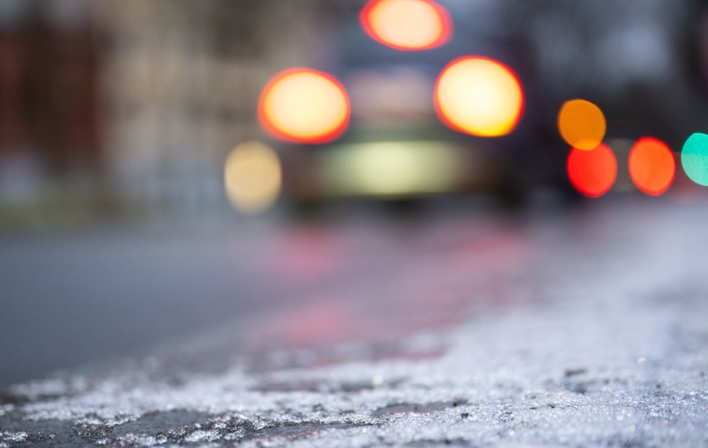 Eine dünne Eisschicht liegt am Morgen auf einem Fahrradweg, während im Hintergrund Autos über eine vereiste Straße fahren.