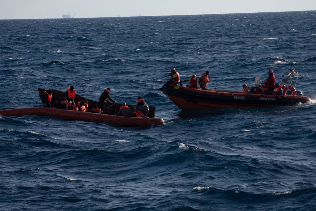 Migranten sitzen in einem Boot im Mittelmeer; Rettungskräfte versuchen, ihnen zu helfen.