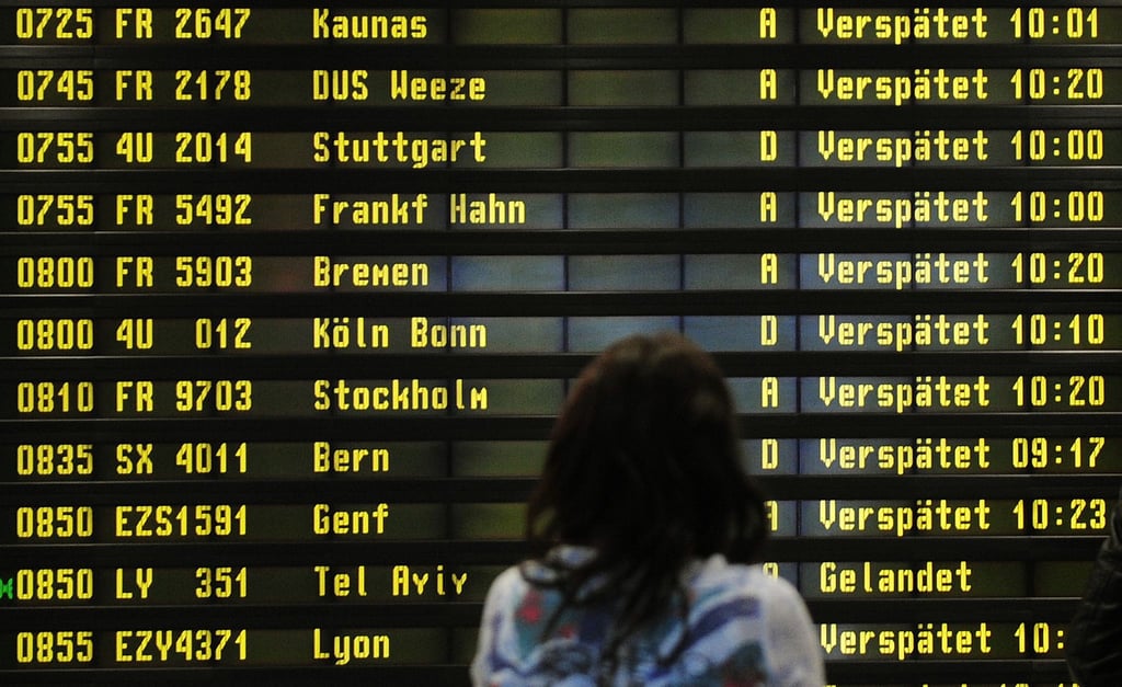 Eine Frau steht auf einem Flughafen vor einer Anzeigetafel, auf der verspätete Flüge aufgelistet werden.