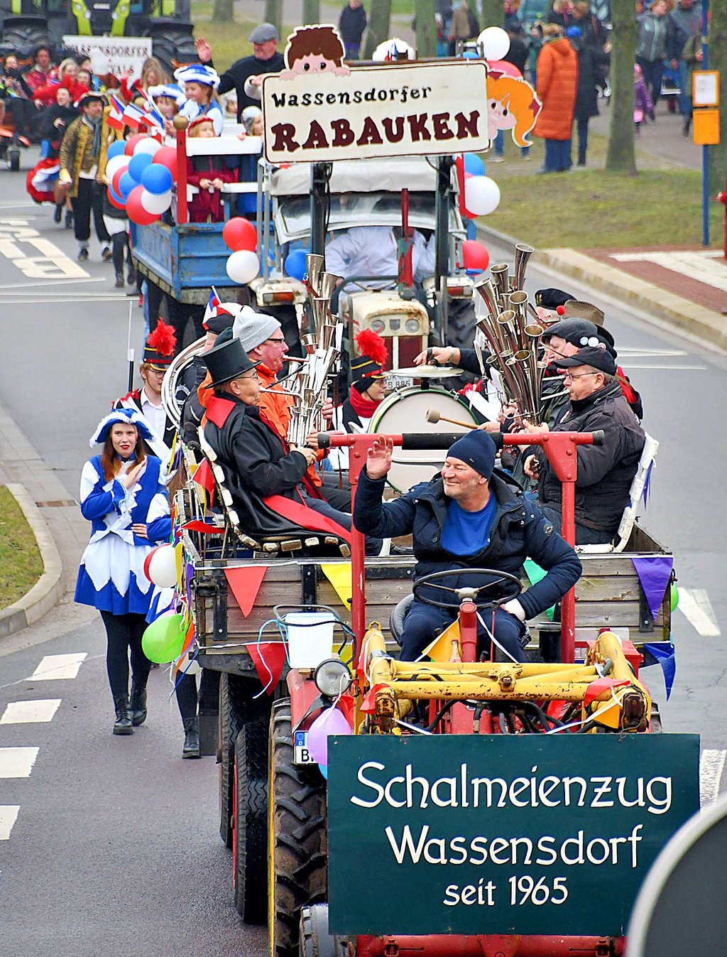 Musiker des Wassensdorfer Schalmeienzuges sitzen auf dem Anhänger, den Ulf Butzlaff mit dem Traktor zieht. Mit dem Schild „Wassensdorfer Rabauken“ weisen die Karnevalisten auf ihr Bedauern hin, dass der Kindergarten geschlossen wurde.