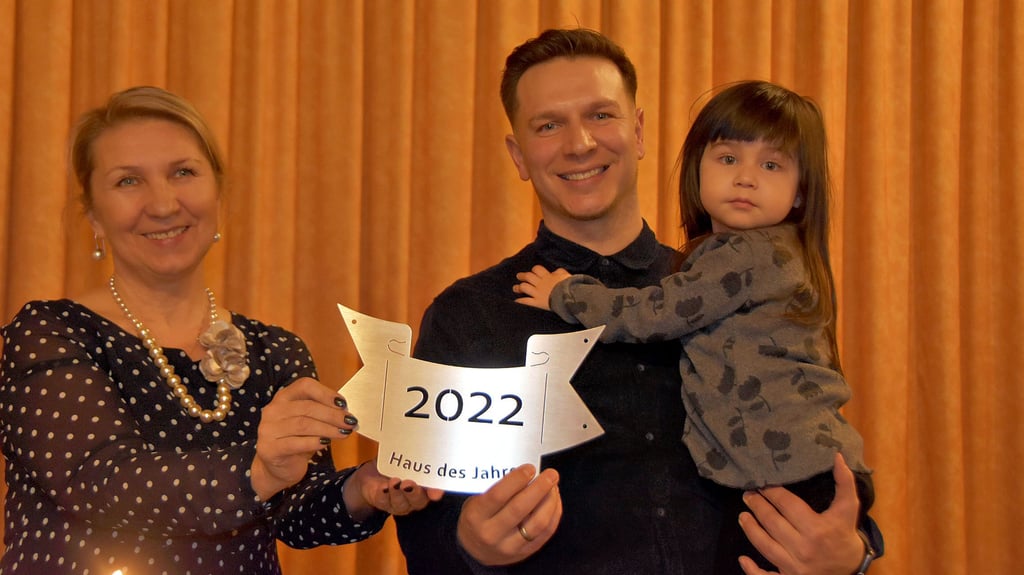 Oebisfeldes Ortsbürgermeisterin Bogumila Jacksch überreicht eine Plakette für das Haus des Jahres 2022 an Eduard Kraus, der seine Tochter auf dem Arm trägt.
