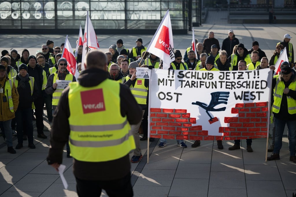 Streikende stehen während einer Verdi–Kundgebung mit einem Banner, auf dem „Schluss jetzt mit Tarifunterschied Ost West“ steht, am Flughafen Berlin Brandenburg.