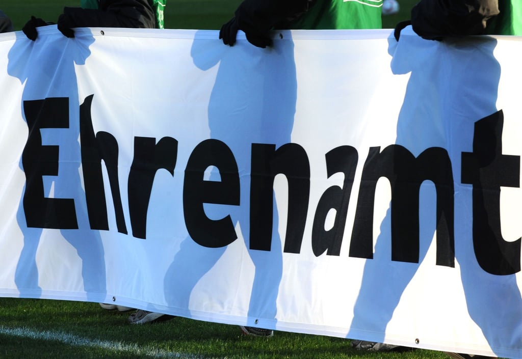 Jugendliche halten vor einem Fußball-Bundesligaspiel ein Banner mit der Aufschrift „Ehrenamt“ in den Händen.